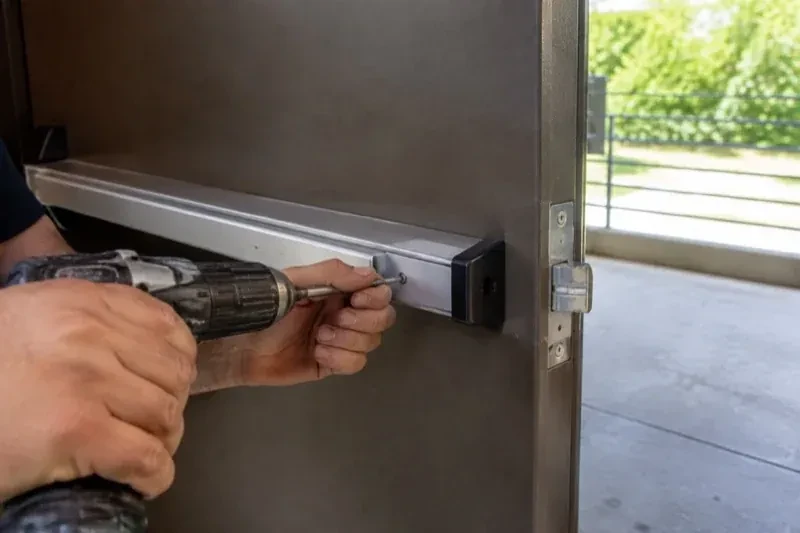 Hands using a power driver at the end of a horizontal push bar on an open metal door. — Panic & Exit Hardware, Kitchener