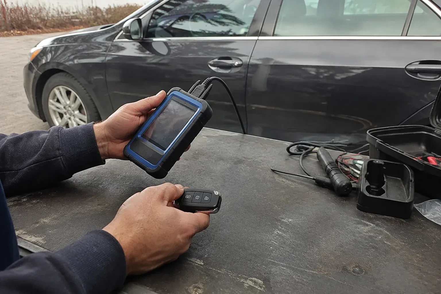 Hands holding a blue-edged electronic device and car-key fob at a worktable beside a parked car. — Transponder Key Programming, Kitchener